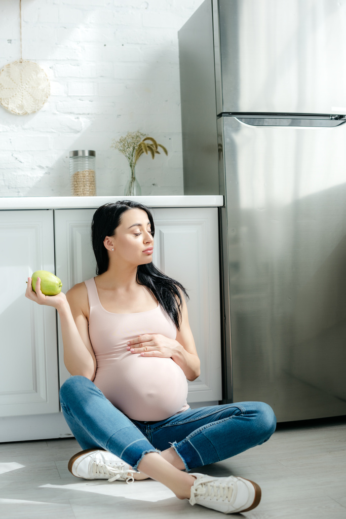 Schwangere hält Apfel auf dem Boden in Küche neben Kühlschrank - Foto, Bild
