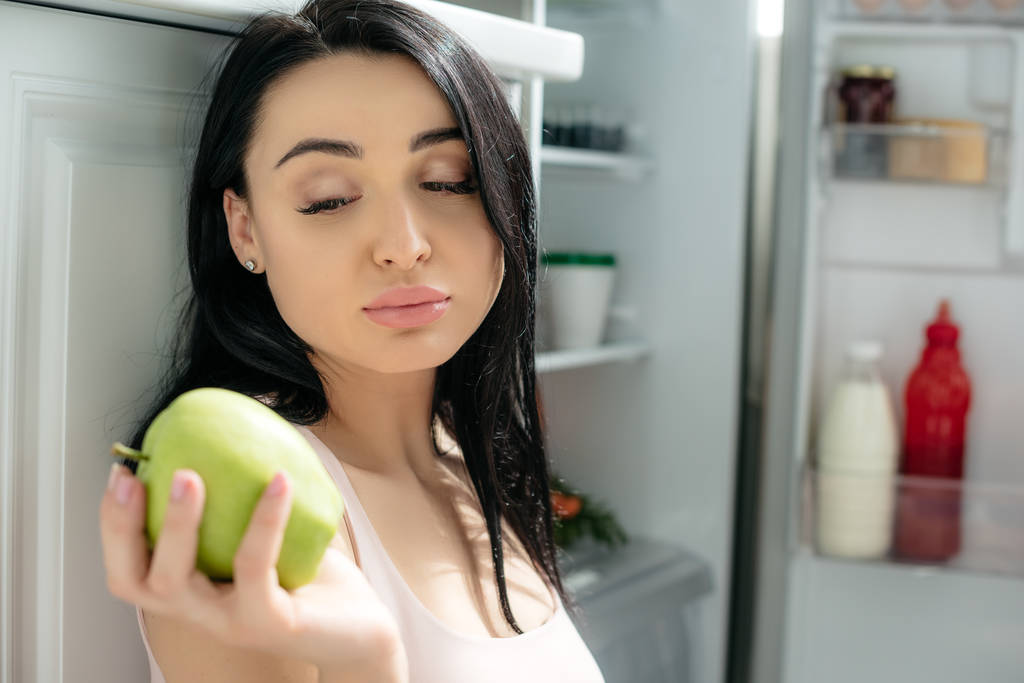 smutná těhotná žena hledá jablko, zatímco sedí poblíž otevřené lednice v kuchyni - Fotografie, Obrázek