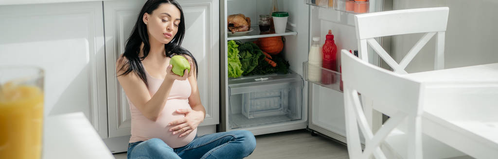 панорамный снимок беременной женщины, держащей яблоко, сидящей на полу на кухне рядом с открытым холодильником
 - Фото, изображение