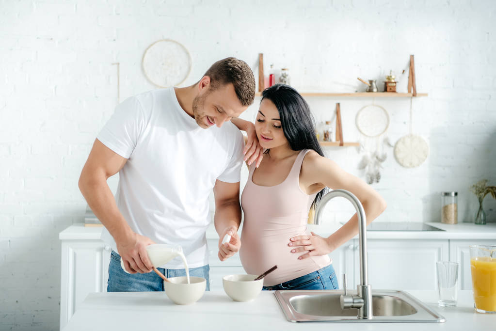 happy pregnant wife and husband making cereals with milk in kitchen - Photo, Image