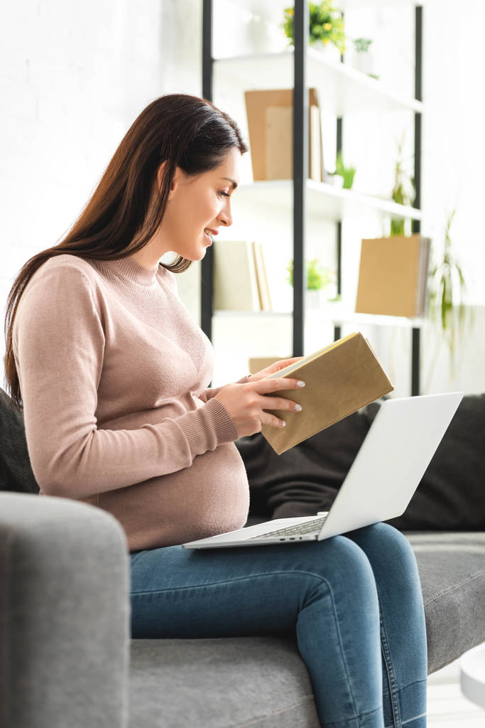 έγκυος γυναίκα κρατώντας βιβλίο, ενώ έχοντας online διαβούλευση με το γιατρό στο φορητό υπολογιστή στο σπίτι  - Φωτογραφία, εικόνα