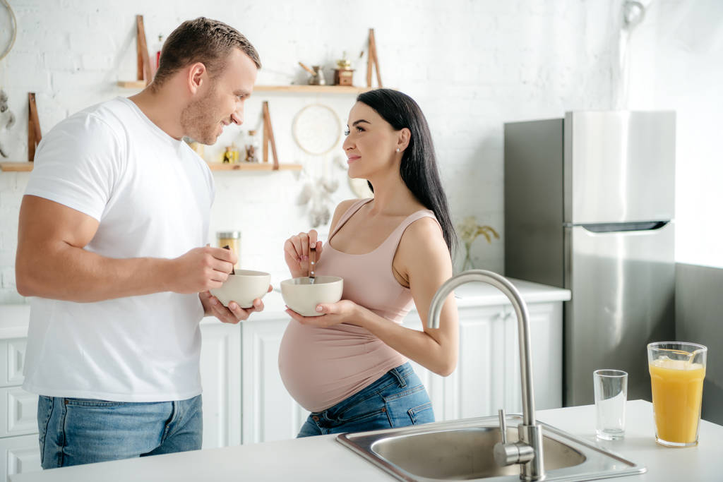 sorrindo casal grávida comer cereais com leite de tigelas na cozinha
 - Foto, Imagem