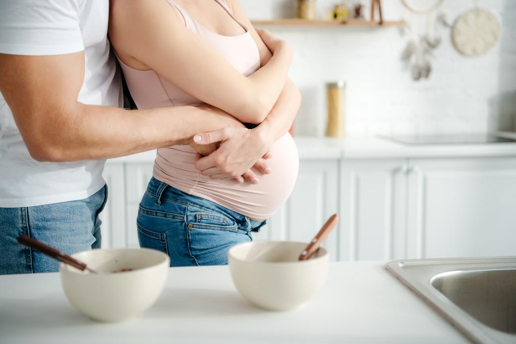 gedeeltelijk uitzicht op zwanger paar knuffelen in de keuken met granen in kommen - Foto, afbeelding
