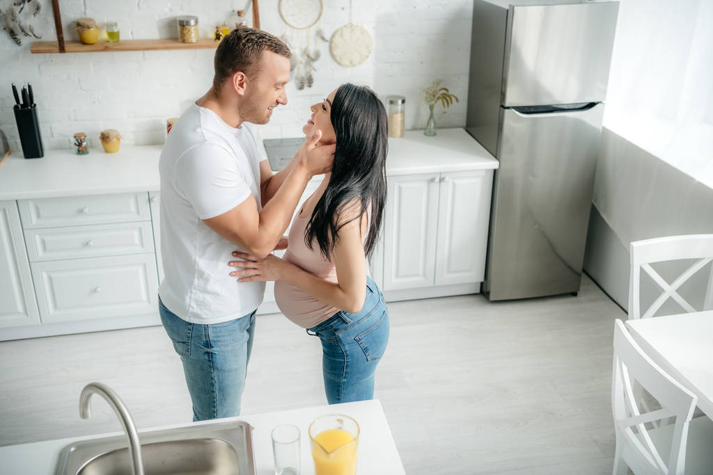 オレンジジュースでキッチンに抱きつく美しい幸せな妊娠中のカップル - 写真・画像