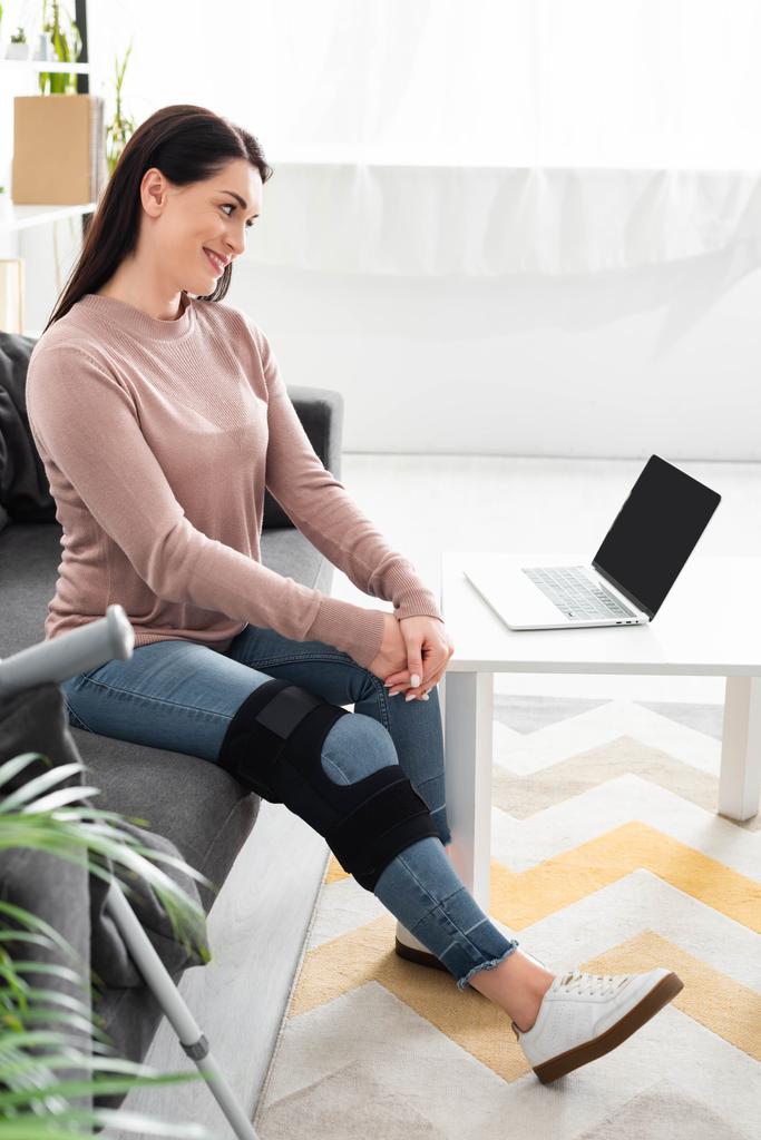 χαμογελαστή γυναίκα με σπασμένο πόδι έχοντας online συμβουλές με το γιατρό στο φορητό υπολογιστή στο σπίτι - Φωτογραφία, εικόνα