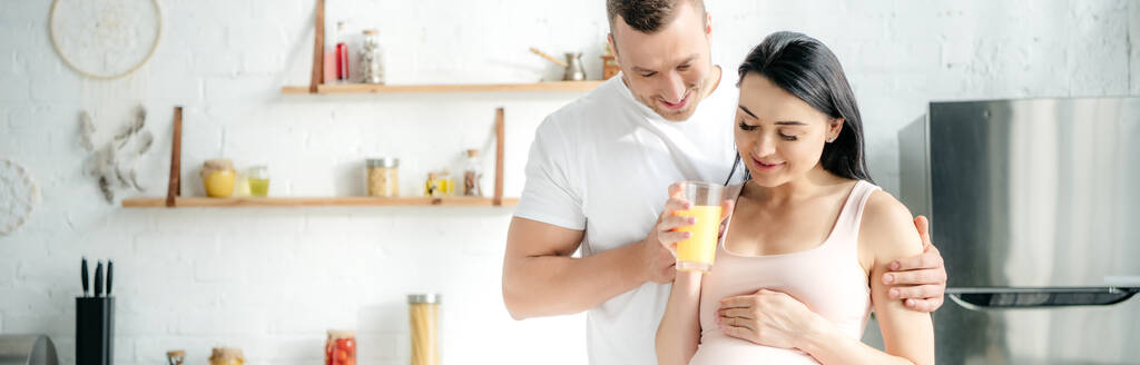 panoramic shot of happy pregnant couple hugging and holding orange juice in kitchen - Photo, Image