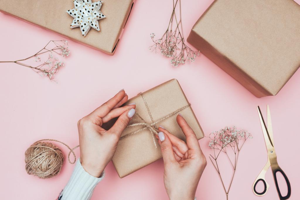 cropped view of girl packing christmas presents with craft paper, twine and flowers, isolated on pink - Photo, Image