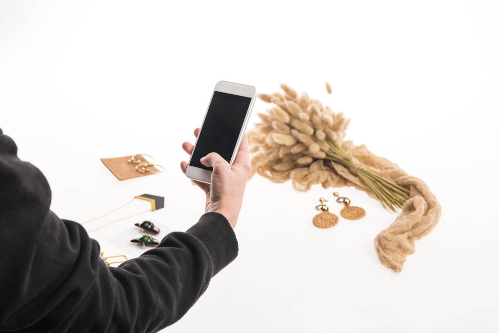 Ausgeschnittene Ansicht eines kommerziellen Fotografen, der Kompositionen mit Flora und Schmuck auf einem Smartphone auf Weiß fotografiert - Foto, Bild