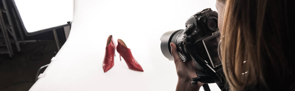 обрізаний вид фотографа, який робить комерційну фотосесію жіночого взуття на червоному каблуці на білому, панорамний знімок
 - Фото, зображення