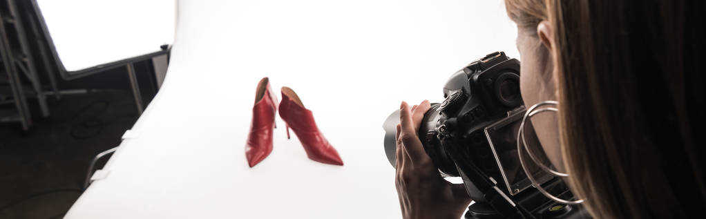 vista recortada de fotógrafo comercial haciendo sesión de fotos comerciales de zapatos de tacón rojo femenino en blanco, tiro panorámico
 - Foto, Imagen