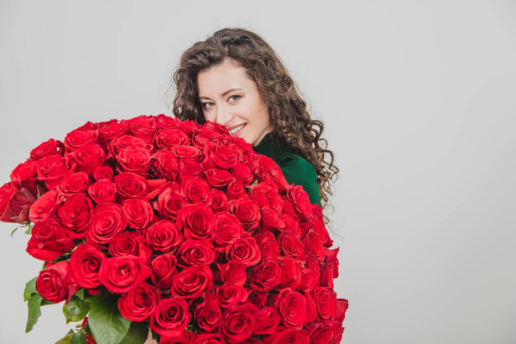 Cute curly brunette girl standing, smelling red roses over white background. - Photo, Image