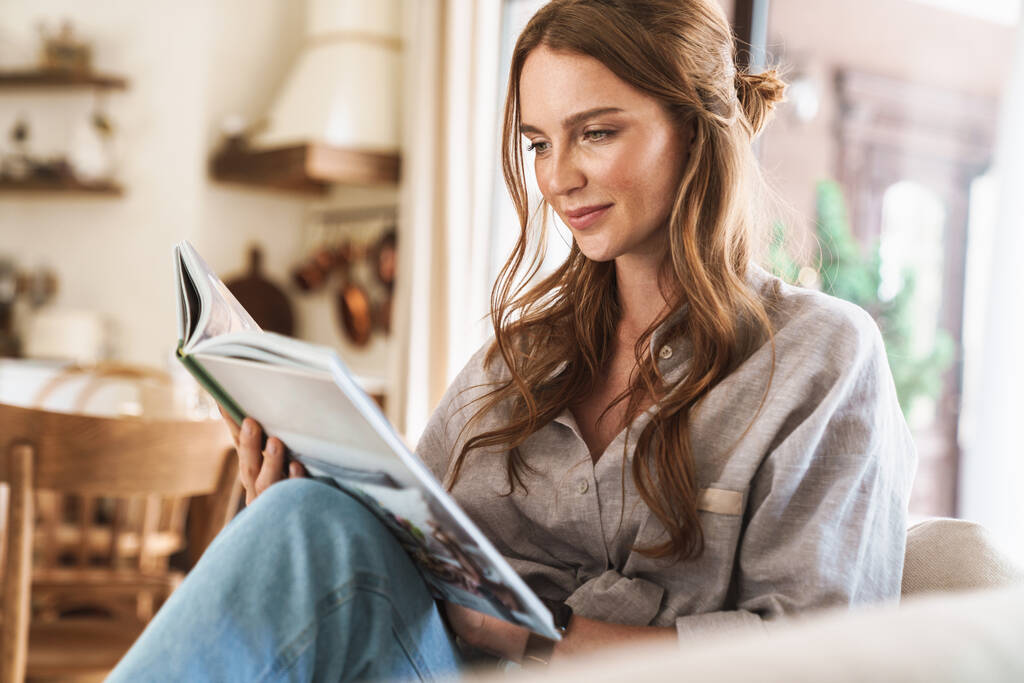 Mujer pelirroja optimista positiva en el interior sentada en casa leyendo libro o revista
. - Foto, Imagen