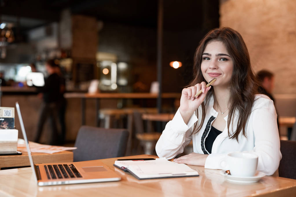 jonge mooie vrouw met pen in haar hand het maken van notities aan haar planner met behulp van haar laptop ziet er tevreden het drinken van hete koffie in cafe moderne zakenvrouw - Foto, afbeelding
