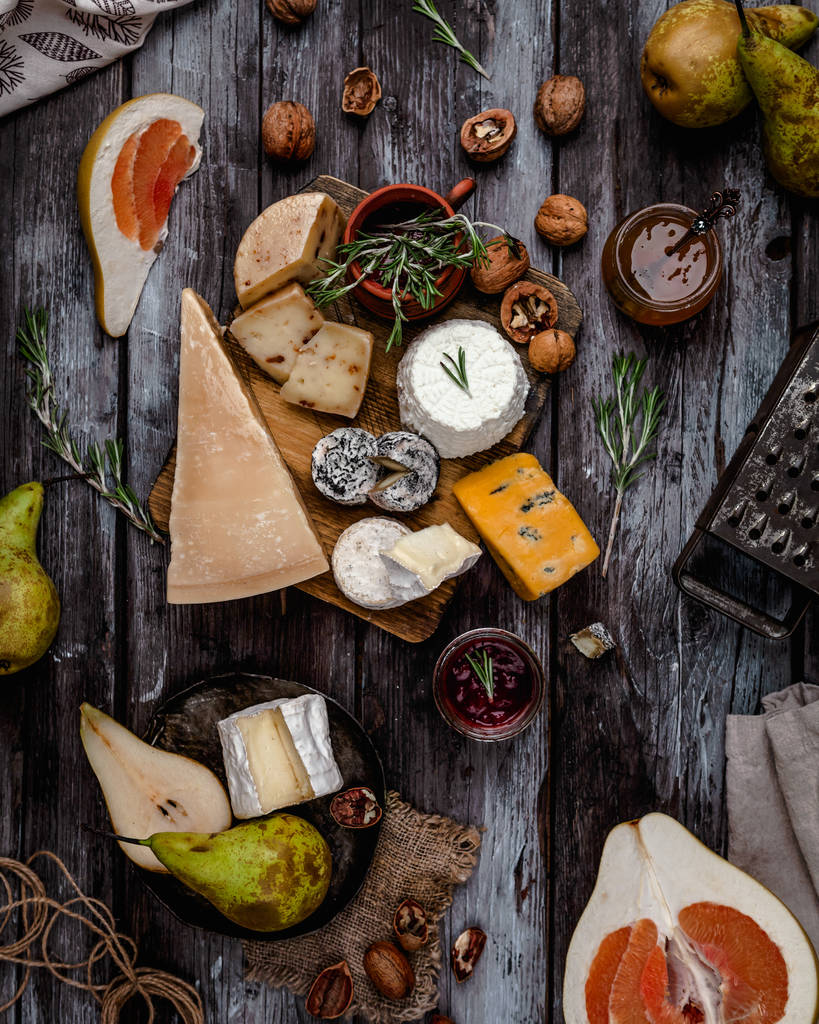Käsesortiment auf Holzbrett mit Honig, Nüssen, Kräutern und Früchten auf braunem Holztisch, mit alter Reibe, Draufsicht - Foto, Bild