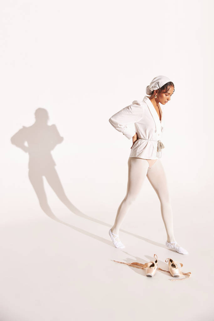 full-length foto van donkergekleurde ballerina op witte achtergrond met schaduw erop, ze draagt wit shirt, beige bodysuit, witte maillot ze staat in de buurt van haar punten en kijkt naar beneden - Foto, afbeelding