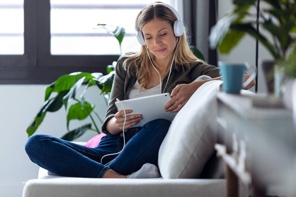 Красивая молодая женщина слушает музыку с наушниками и ее цифровой планшет, сидя на диване дома
. - Фото, изображение