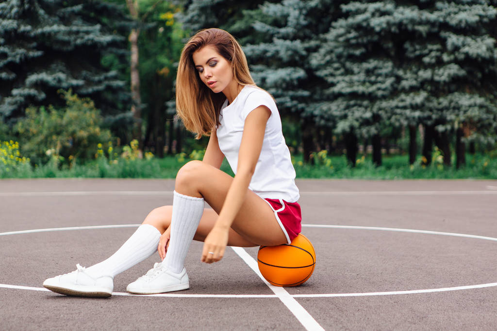 Όμορφη νεαρή κοπέλα ντυμένος με λευκό μπλουζάκι, σορτς και αθλητικά παπούτσια, κάθεται σε ένα γήπεδο μπάσκετ στην μπάλα. - Φωτογραφία, εικόνα