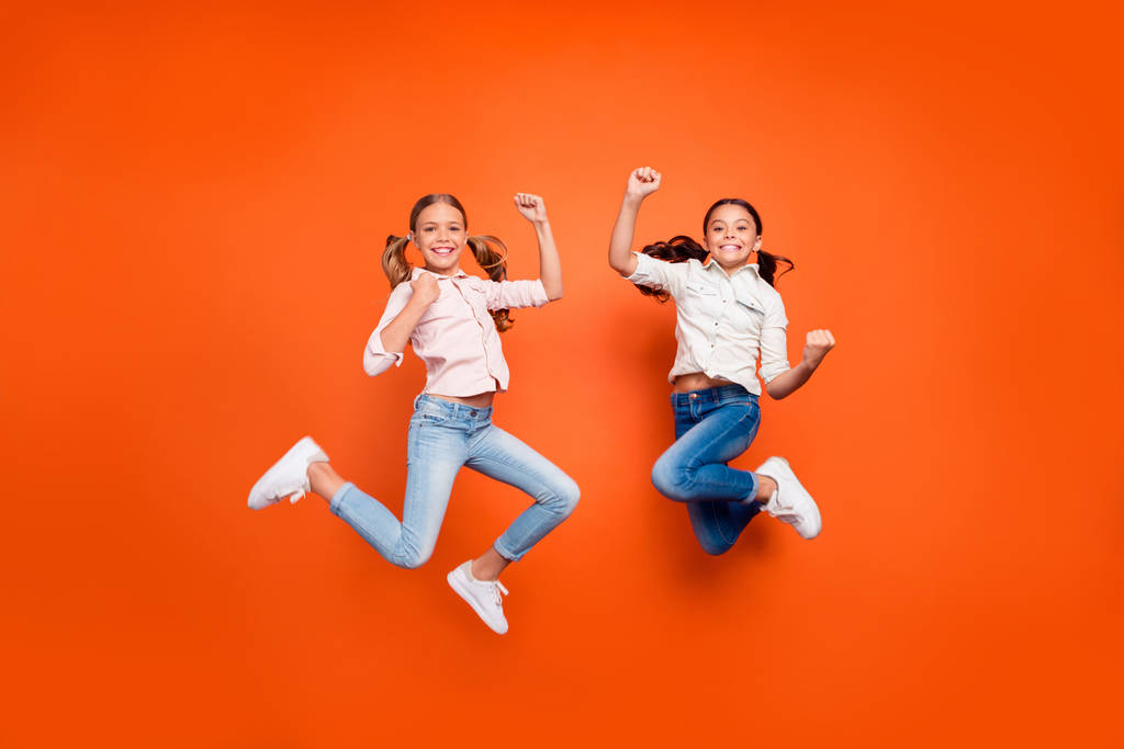 Foto de longitud completa de positivo lindo dos niñas salto celebrar la victoria en el concurso escolar levantar los puños muestran fuerza desgaste camisa blanca vaqueros aislados fondo de color naranja
 - Foto, imagen