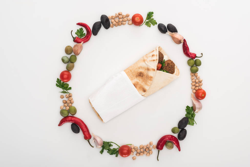 vrchní pohled na cizrnu, česnek, cherry rajčata, petržel, olivy, chilli paprika, zelená cibule uspořádány v kulatém rámu kolem falafel v pita na bílém pozadí - Fotografie, Obrázek