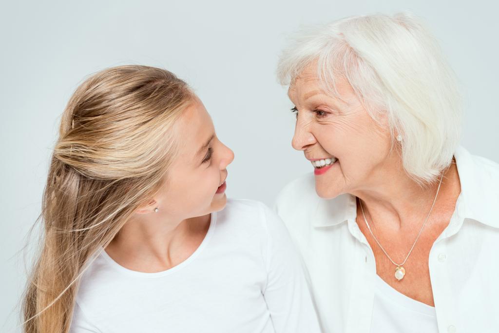 uśmiechnięta wnuczka i babcia patrzące na siebie na szarym  - Zdjęcie, obraz
