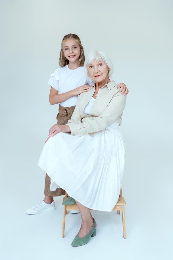 Lächelnde Enkelin umarmt Großmutter auf Hocker vor grauem Hintergrund  - Foto, Bild