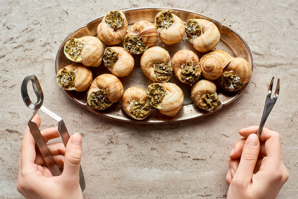 partial view of woman eating delicious cooked escargots with tweezers on stone background - Photo, Image
