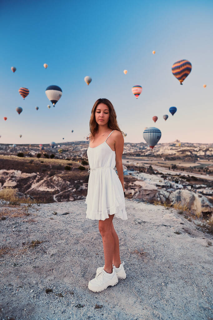 Een toeristisch meisje met een witte trui op een bergtop met een prachtig uitzicht op ballonnen in Cappadocië. Happy Travel in Turkije concept - Foto, afbeelding