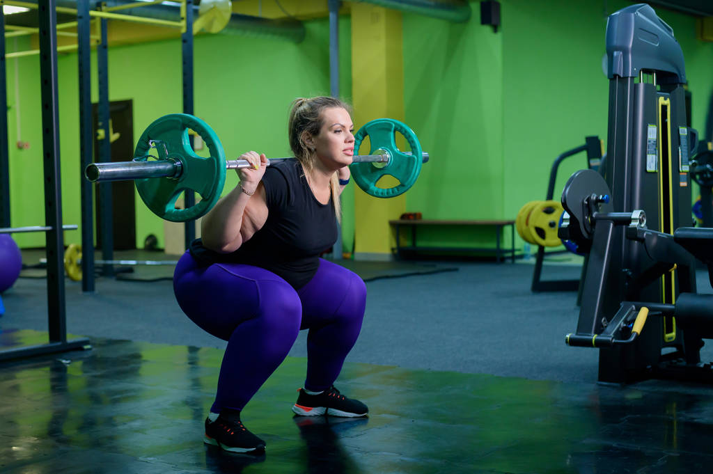 Η υπέρβαρη γυναίκα σκύβει με ένα κουδούνι στο γυμναστήριο. Ένα χοντρό κορίτσι κάνει ασκήσεις στους γλουτούς. Πηγαίνει για σπορ για την απώλεια βάρους. - Φωτογραφία, εικόνα