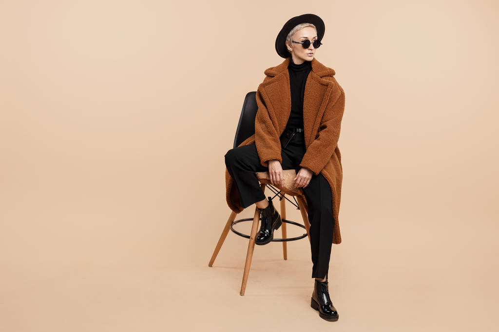 Serious young hipster woman with blonde short hair wearing a coat, hat and sunglasses posing over beige background. - Photo, Image