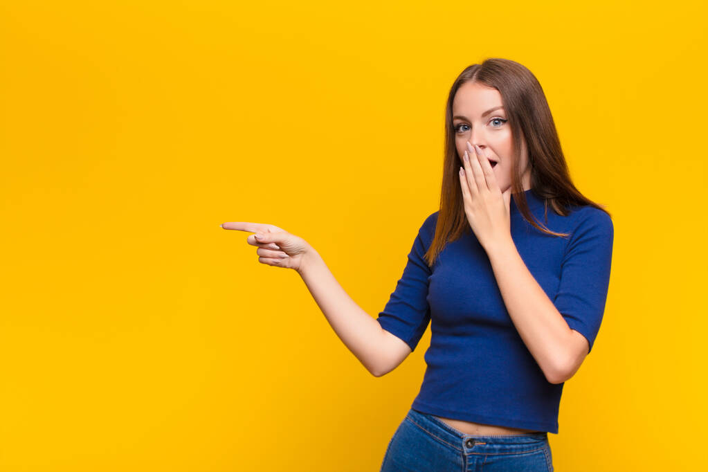 young red head woman feeling happy, shocked and surprised, covering mouth with hand and pointing to lateral copy space against flat wall - Photo, Image
