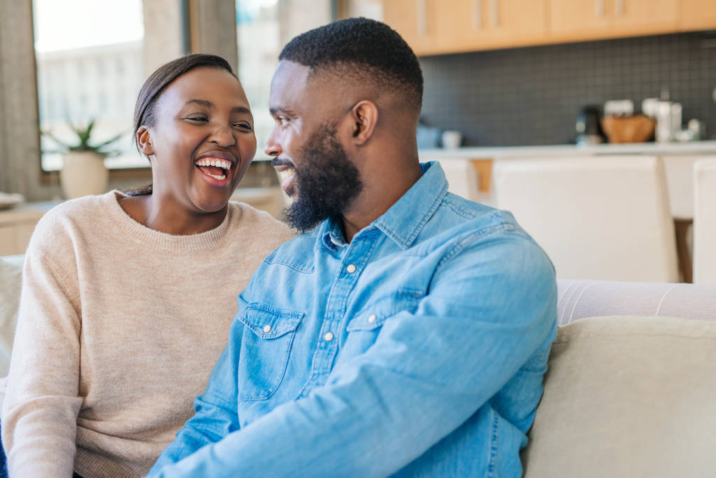 Mladá Afroameričanka se směje, zatímco sedí na pohovce v obývacím pokoji doma se svým manželem - Fotografie, Obrázek
