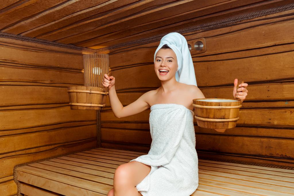 glimlachende en aantrekkelijke vrouw in handdoeken met wastafels in de sauna  - Foto, afbeelding