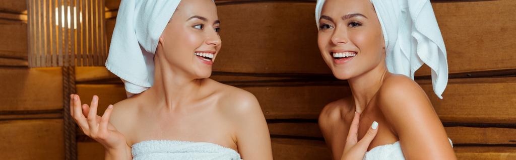 panoramiczne ujęcie uśmiechniętych i atrakcyjnych przyjaciół w ręcznikach rozmawiających w saunie  - Zdjęcie, obraz