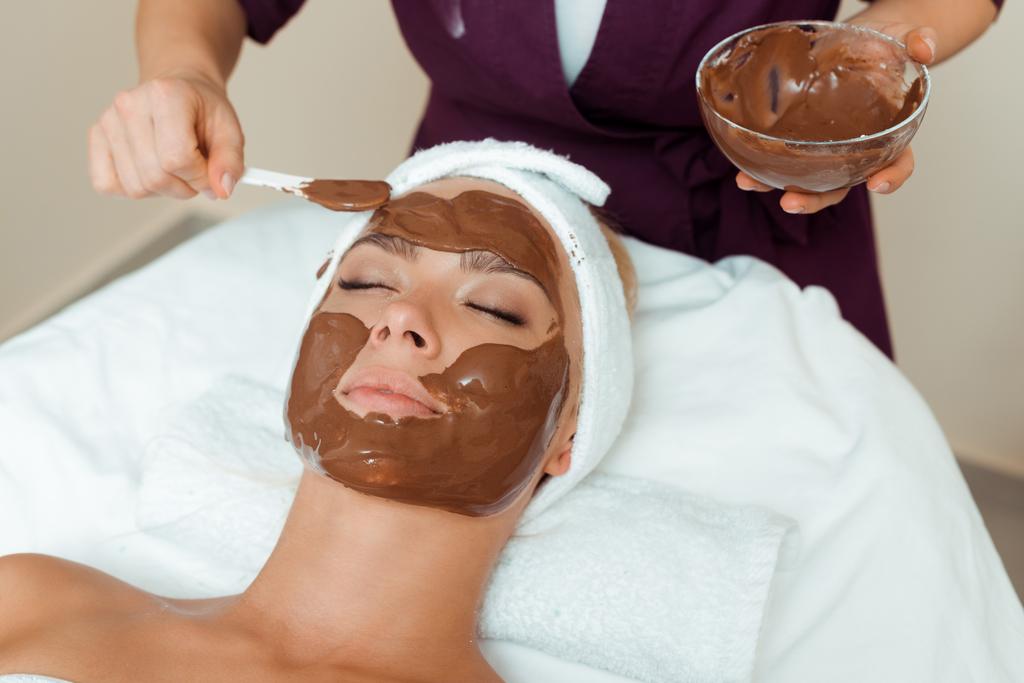 обрізаний вид косметолога, що наносить маску для обличчя на привабливу жінку в спа
  - Фото, зображення