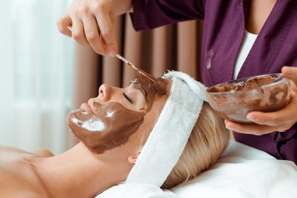 Kozmetik uzmanının kaplıcadaki çekici kadına yüz maskesi takması.  - Fotoğraf, Görsel