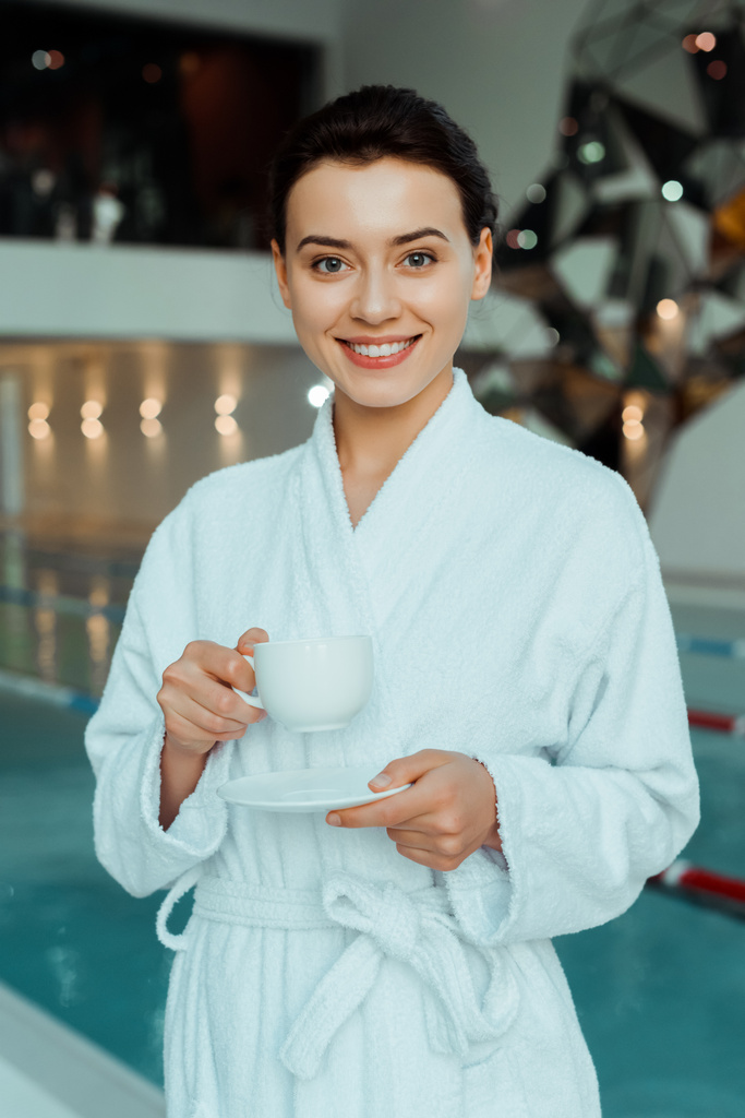 femme attrayante et souriante en peignoir blanc tenant tasse de café dans le spa
  - Photo, image