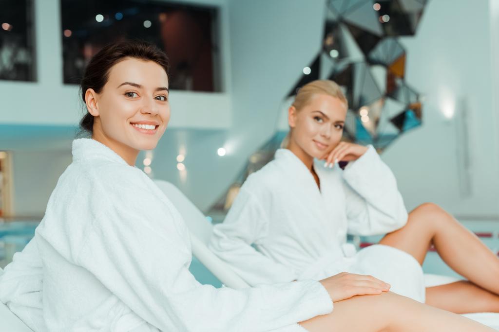 selective focus of attractive and smiling woman looking at camera and friend on background in spa  - Photo, Image