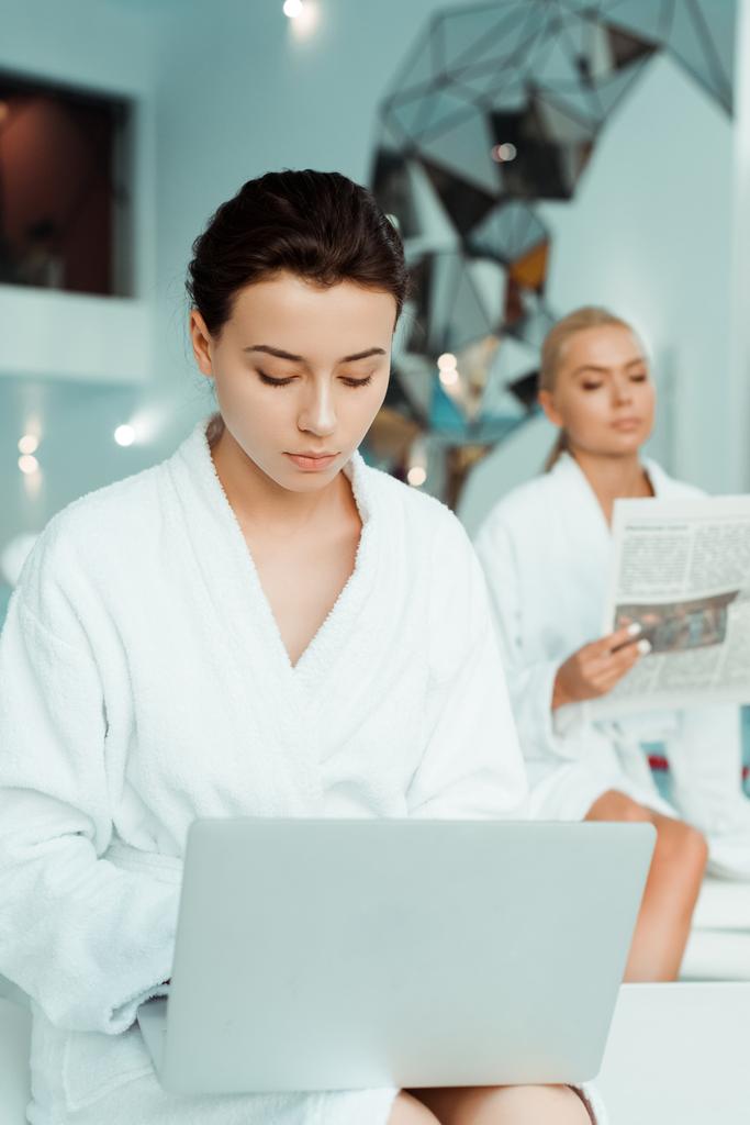 επιλεκτική εστίαση ελκυστική γυναίκα χρησιμοποιώντας φορητό υπολογιστή και φίλο ανάγνωση εφημερίδα στο παρασκήνιο σε spa  - Φωτογραφία, εικόνα