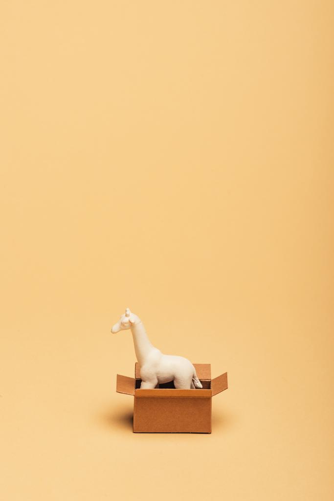 White toy giraffe in cardboard box on yellow background, animal welfare concept - Photo, Image