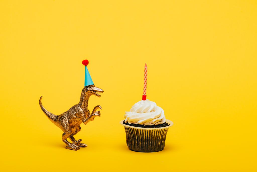 Dinosauro giocattolo in cappuccio partito e cupcake con candela su sfondo giallo
 - Foto, immagini