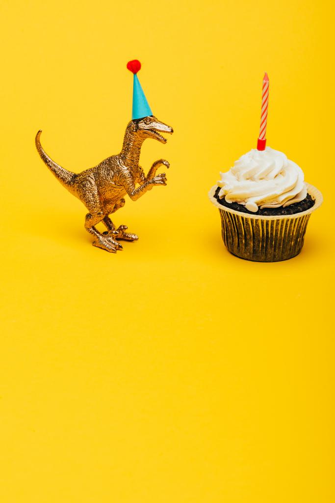 Toy dinosaur in party cap beside cupcake with candle on yellow background - Photo, Image