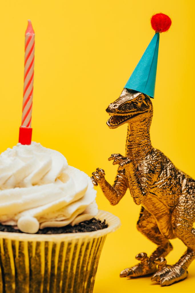 Foco selectivo de dinosaurio de juguete en gorra de fiesta junto a magdalena con vela sobre fondo amarillo
 - Foto, imagen