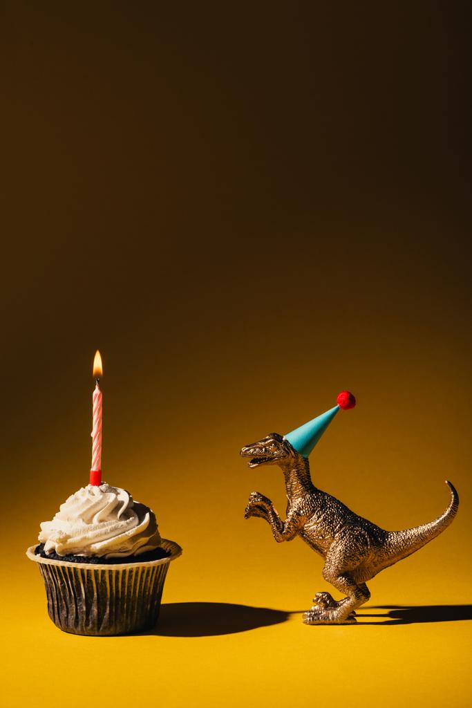 Speelgoeddinosaurus in party cap naast cupcake met brandende kaars op bruine achtergrond - Foto, afbeelding