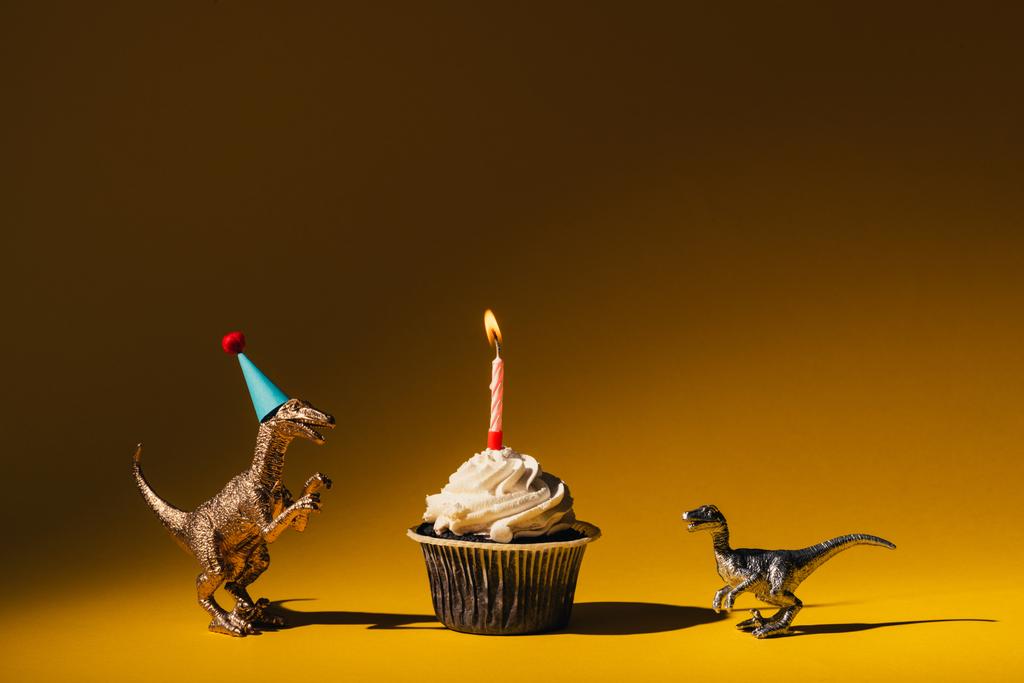 Parti şapkalı oyuncak dinozorlar ve turuncu arka planda yanan mumlu kek. - Fotoğraf, Görsel
