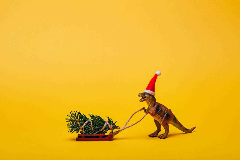 Toy dinosaur in santa hat with pine on sleigh on yellow background - Photo, Image