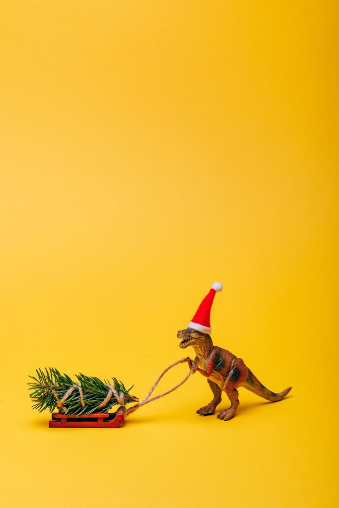 Παιχνίδι δεινόσαυρος στο καπέλο santa με έλατο στο έλκηθρο σε κίτρινο φόντο - Φωτογραφία, εικόνα