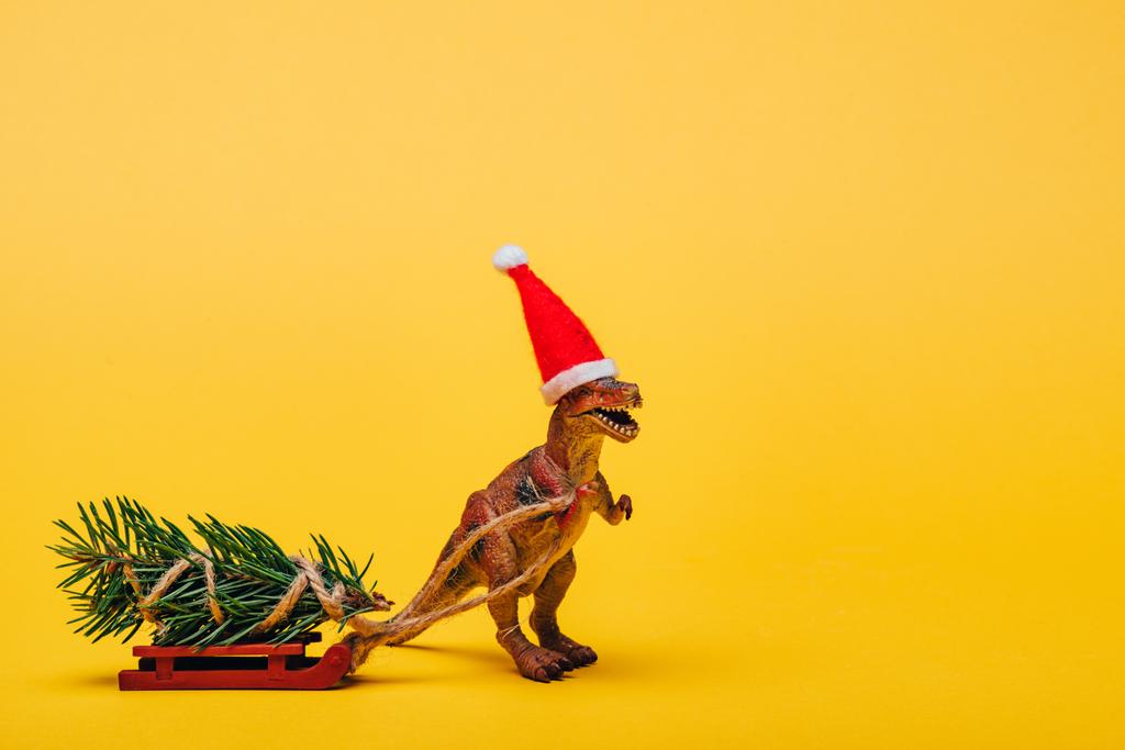 Zabawkowy dinozaur w kapeluszu Santa z jodłą na saniach na żółtym tle - Zdjęcie, obraz