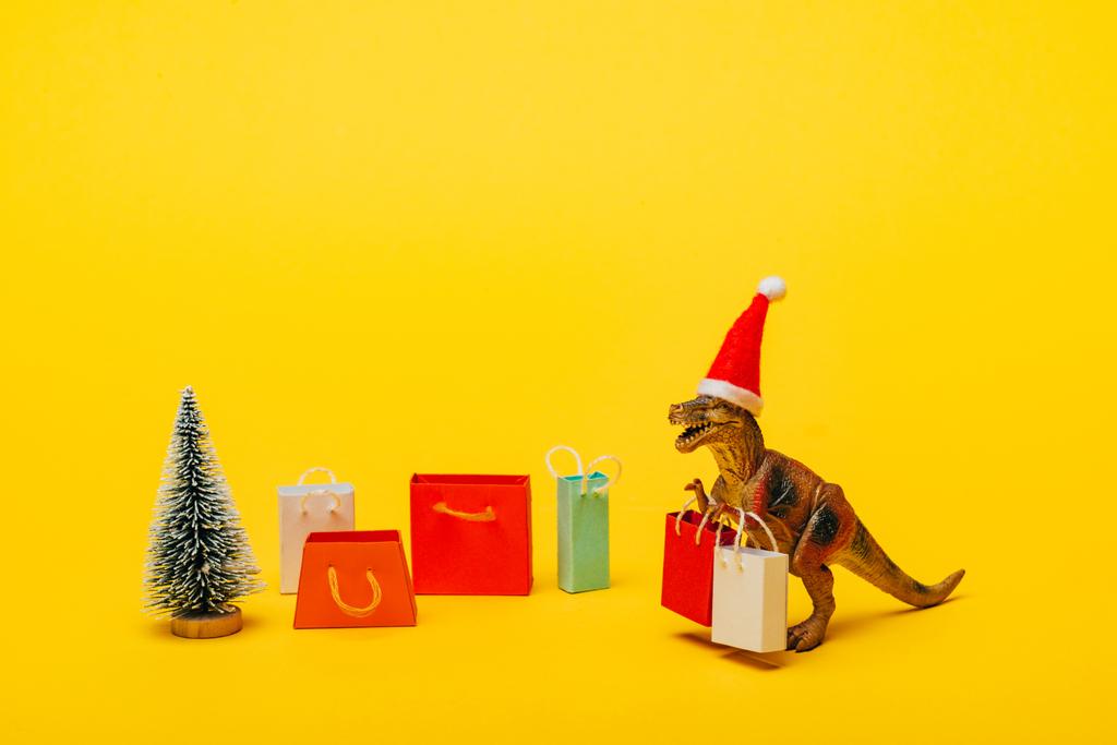 Toy dinosaur in santa hat with shopping bags and christmas tree on yellow background - Photo, Image