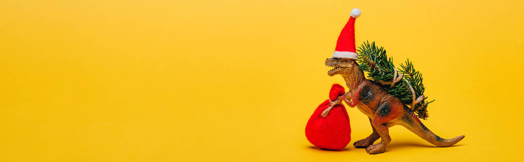Dinosauro giocattolo in cappello di Babbo Natale con sacco e abete su sfondo giallo, scatto panoramico
 - Foto, immagini