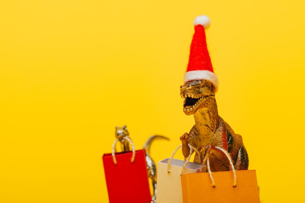 Enfoque selectivo de dinosaurios de juguete en sombrero de santa y bolsas de compras sobre fondo amarillo
 - Foto, imagen